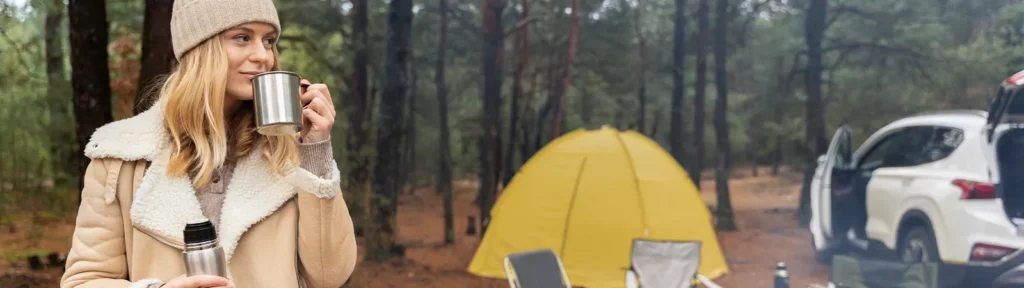 Campingplaetze Deutschland - Frau steht im Wald mit einer Thermoskanne und hinter ihr steht das aufgebaute Camping-Zelt
