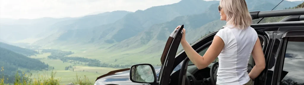 Car-Rental-Service - Frau steht neben einem schwarzen Auto und schaut eine bergige Landschaft an