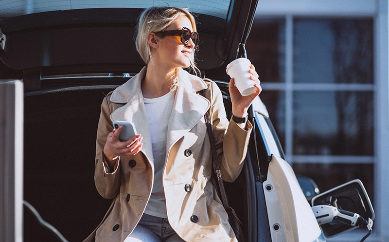 Reise mit E-Auto planen - Frau sitzt mit Kaffee und Handy in der Hand auf dem Kofferraum ihres E-Autos, welches gerade an einer Ladestation aufgeladen wird